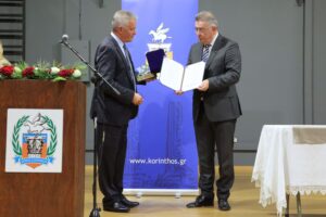 Two men are exchanging an award or certificate at a formal event, with a podium and a decorative backdrop.