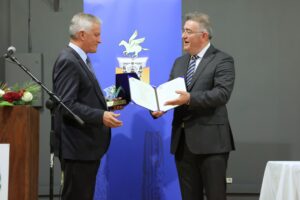 Two men are exchanging an award or certificate at a formal event, with a blue banner in the background.
