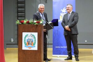 Two men are smiling and standing near a podium with flowers, engaged in a conversation during an event.