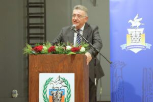 A man in a suit speaks at a podium decorated with flowers, with a blue backdrop featuring a logo.