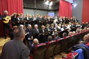 A choir performs on stage, accompanied by a guitarist, while an audience watches from red seats. Red curtains frame the scene.