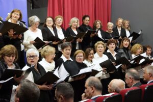A choir of women in formal attire performs on stage, holding music sheets, with an audience visible in front.