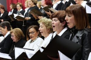 A choir of women singing together, holding music sheets, dressed in black and white attire.