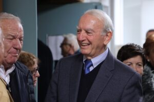 Two older men are engaged in conversation, with one smiling warmly. A small group of people is visible in the background.