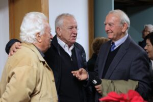Three older men are engaged in conversation, smiling and gesturing, in a warmly lit indoor setting.