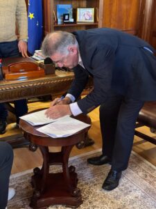 A man in a suit is signing documents on a small table in an office setting, with a European flag in the background.