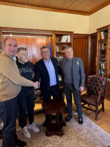 Four individuals are shaking hands in a warmly decorated office, with bookshelves and a wooden desk in the background.