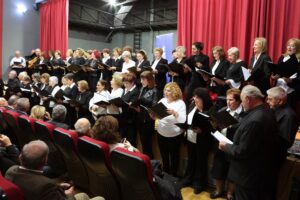 A large choir of women in black and white attire performs on stage, with an audience seated in front. Red curtains frame the scene.