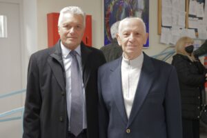 Two men stand side by side, both dressed in formal attire, smiling at the camera in a brightly lit indoor setting.