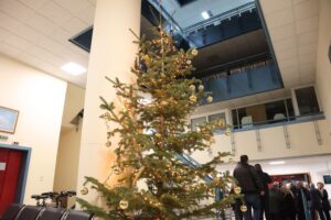 A decorated Christmas tree stands in a spacious indoor area, with people gathered in the background.