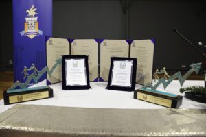 A display of awards on a table, featuring two framed certificates and three decorative trophies, with a backdrop of a logo.