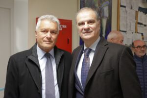 Two men in formal attire pose for a photo in an indoor setting, with a red cabinet and people in the background.