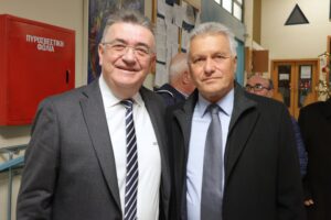 Two men in formal attire pose for a photo, smiling, in an indoor setting with a fire safety box in the background.