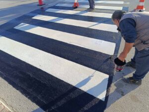 A worker paints a black and white crosswalk, with traffic cones nearby.