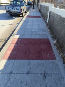 A sidewalk with alternating red and gray tiles, alongside a street with parked vehicles and a wall.