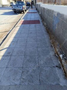 A sidewalk with textured concrete, featuring red and gray tiles, alongside a street with parked vehicles.