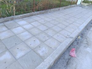 A newly paved concrete sidewalk with a grid pattern, bordered by a low wall and some litter on the side.
