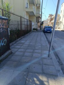 A sidewalk lined with buildings and a parked blue car, featuring a fence and some greenery on the left.