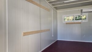 An empty room with light gray walls, wooden strips for hanging, and a window. The floor is dark, and the ceiling is white.