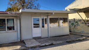 A small, modern building with a white exterior and large windows, situated on a street with graffiti on the adjacent wall.