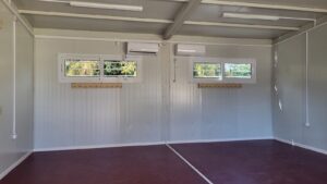 An empty interior of a room with gray walls, two windows, air conditioning units, and a maroon floor.