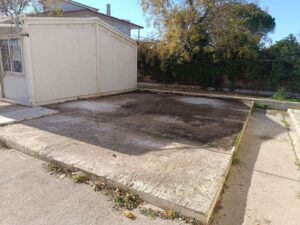 A vacant, concrete area with some dirt patches, adjacent to a small white structure and surrounded by greenery.