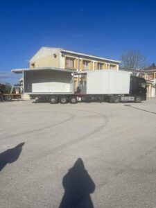 A large truck with a modular structure is parked in front of a building on a sunny day.